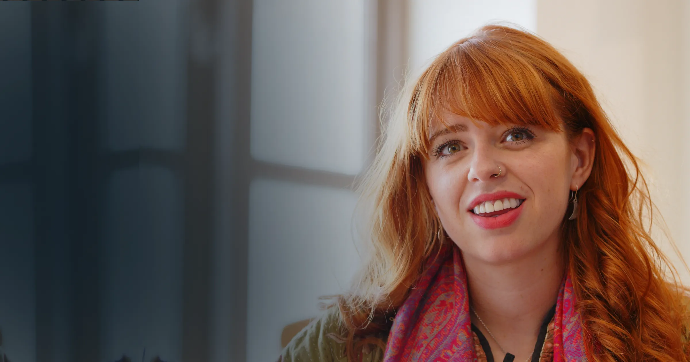 A woman with red hair smiles warmly, wearing a colourful scarf. The background is softly blurred, suggesting an indoor setting with natural light.